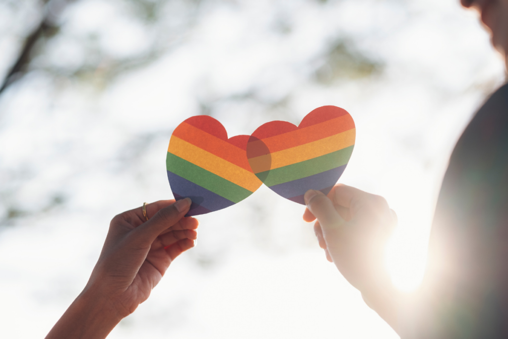 two people holding up paper hearts with the the pride flag in them in front of a clear sky